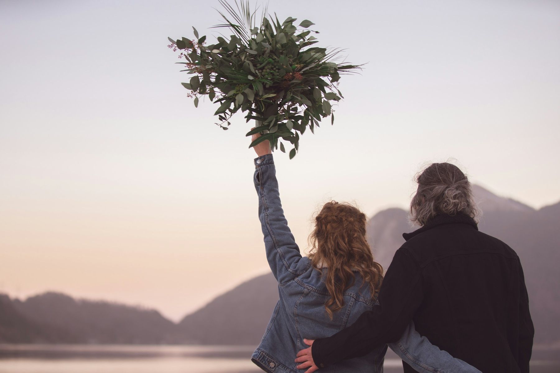 Un couple de dos face au lac au crépuscule, elle lève un bouquet de feuillage vers le ciel