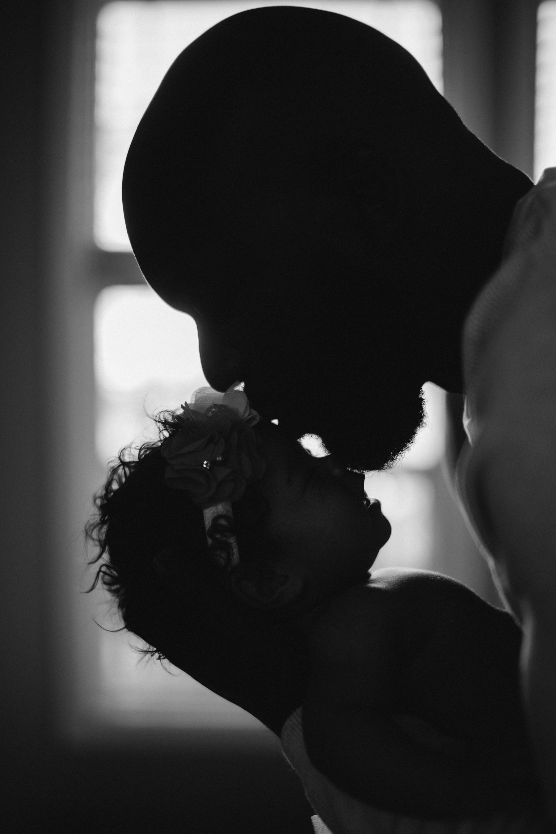 En noir et blanc, une maman embrasse son bébé près d’une fenêtre